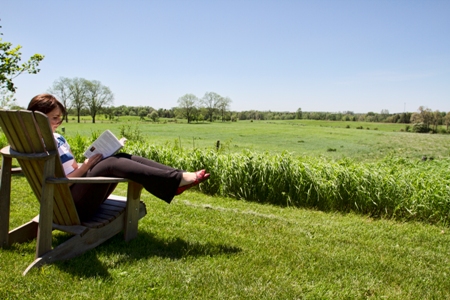 Photo of a young woman reading a book in a wooden chair by a green field in summer